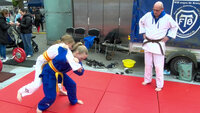 Zwei Mädchen führen eine Judo-Wurftechnik auf einer roten Matte vor, während ein Trainer mit braunem Gürtel aufmerksam zuschaut. Die Szene findet im Freien auf dem Vegesacker Kindertag statt.